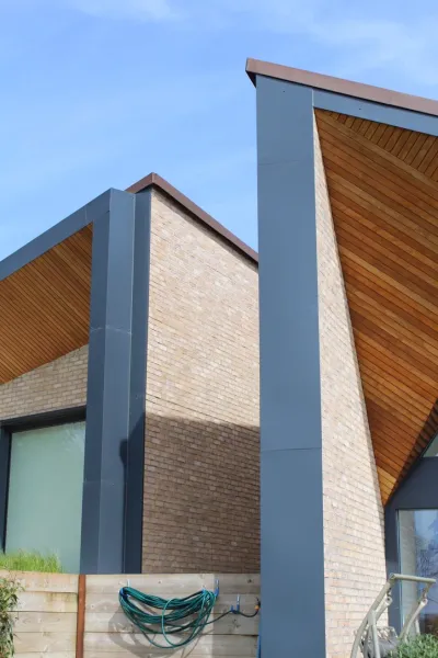 Roofline detail at Weald House, where warm timber-clad eaves meet brickwork and a dark zinc fascia against a blue sky.