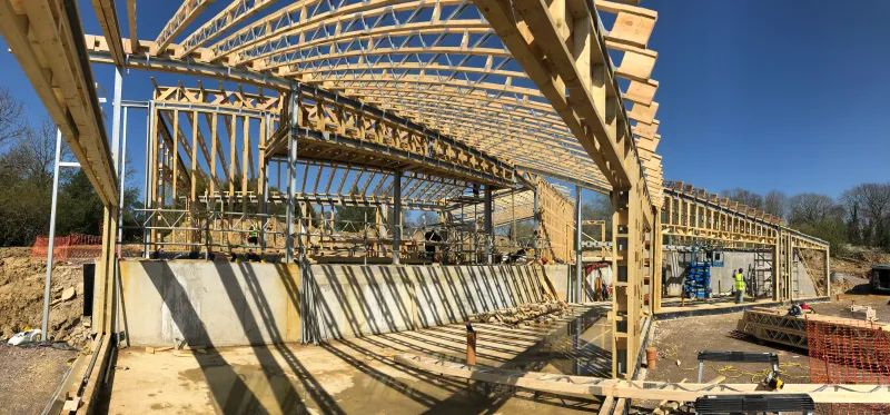 A timber-frame new build under construction, with engineered roof trusses and a stud frame rising over concrete foundations.