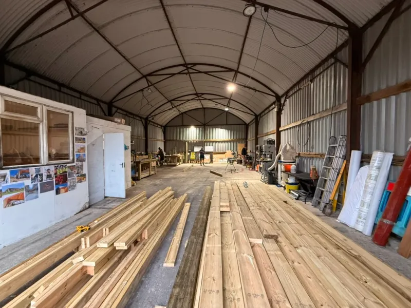 Inside Tidal Drift's workshop: a curved-roof barn with a steel portal frame, prepared timber stacked on the floor and benches and machinery along the far wall.