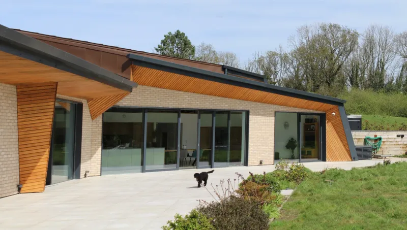 A completed contemporary single-storey new build with a sweeping timber-lined roof, buff brickwork and full-height sliding glazing, set in open countryside.