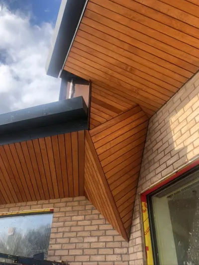 Looking up at precision timber cladding meeting at a mitred corner, with a dark zinc fascia over buff brickwork against a cloudy sky.