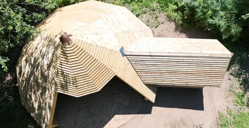 The finished ten-sided fire-pit hut from above, its timber roof rising to a point beside a boarded store, in a woodland clearing.