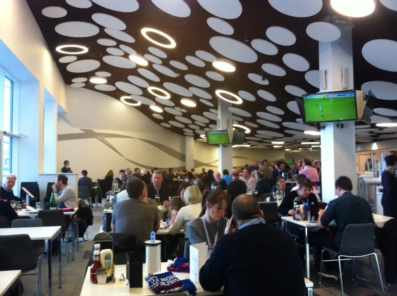 The completed stadium hospitality lounge in use, busy with guests at tables under the circular-disc ceiling, with screens showing the match.