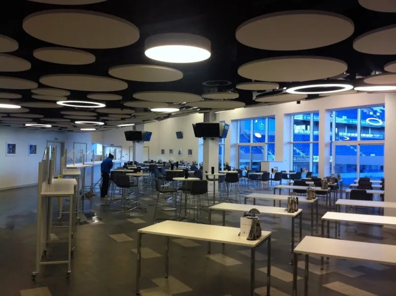 A stadium hospitality lounge with café tables and bar stools beneath the circular-disc ceiling, with full-height windows overlooking the pitch at dusk.