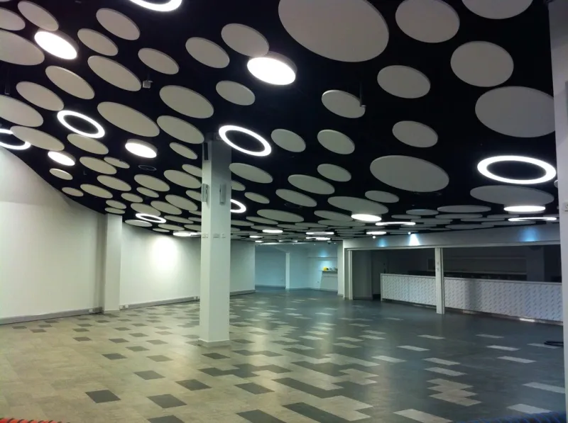 A stadium hospitality concourse with a sculptural black ceiling of white circular acoustic discs and ring lights, above a chequerboard tiled floor.