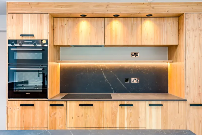 Timber kitchen units with lit shelves above a dark marbled splashback and built-in ovens