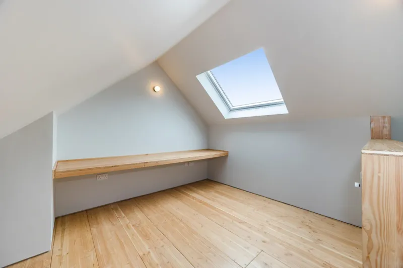 A timber desk built along the wall of a sloping-ceilinged room with a roof light and timber floor