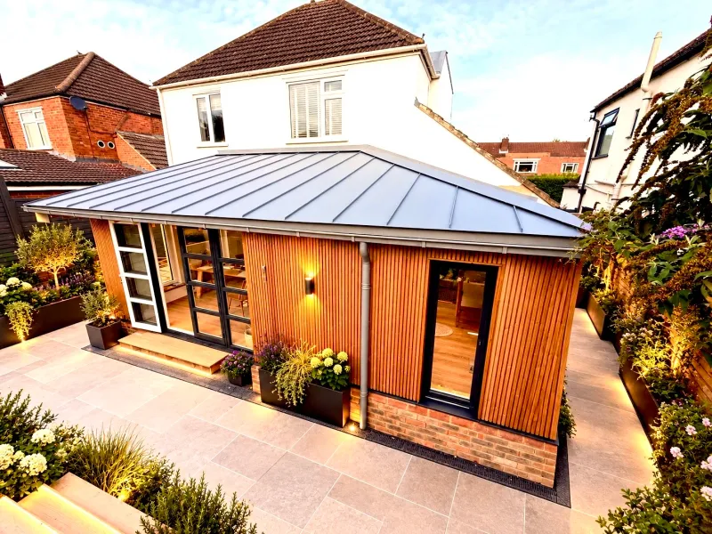 The extension's hipped zinc roof and redwood walls from above at dusk, with the lit terrace and planting around it.