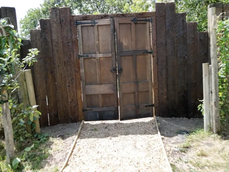 Heavy timber entrance gates set in a wall of upright sleepers, opening onto a gravel path into the woodland.