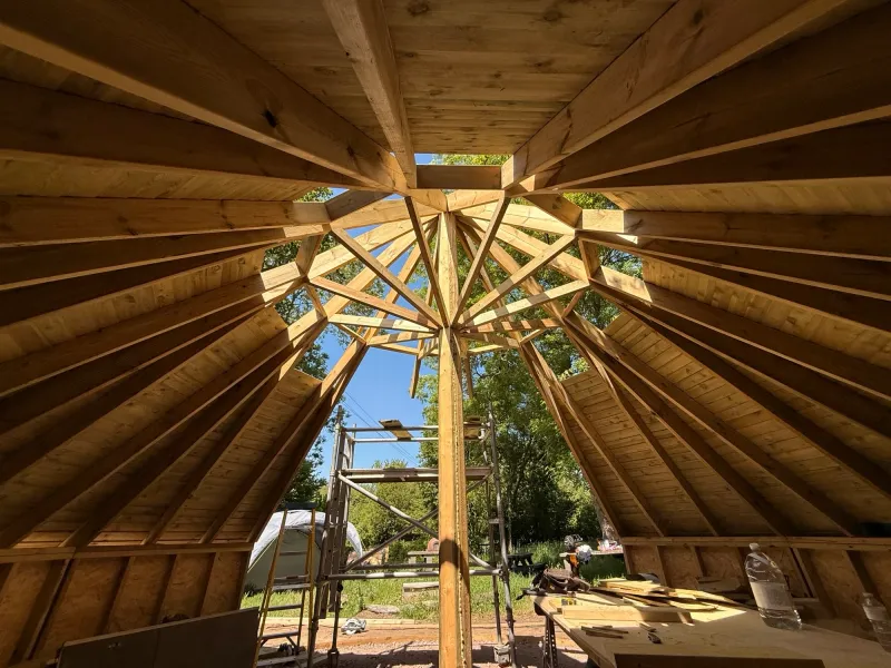 Inside the fire-pit hut during the build, looking up: timber rafters radiating to a ring at the apex of the roof.