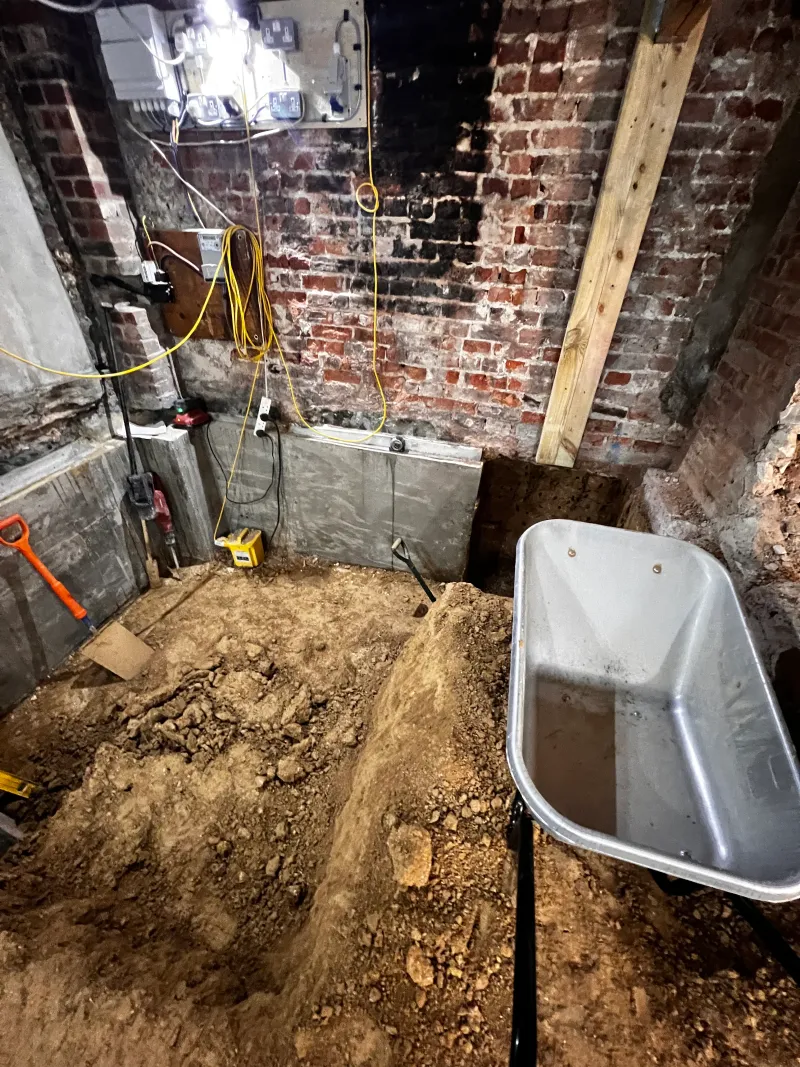 The basement dug down by hand to bare earth, with a wheelbarrow and new underpinning along the wall