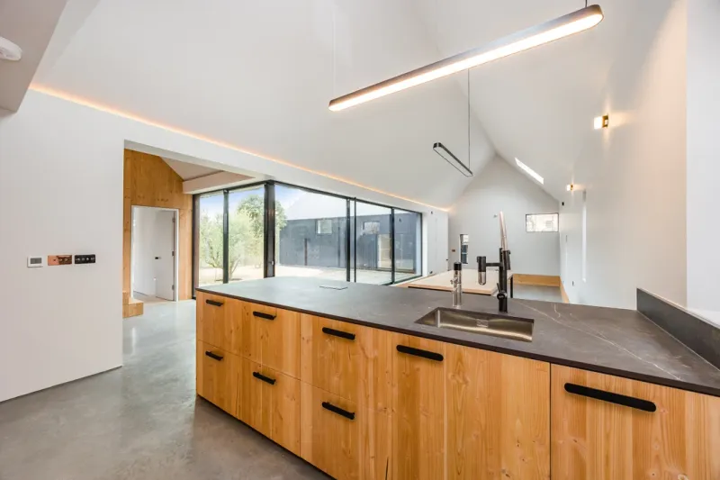 A finished kitchen with a bespoke timber island, dark stone worktop and a vaulted ceiling lit by a linear pendant.