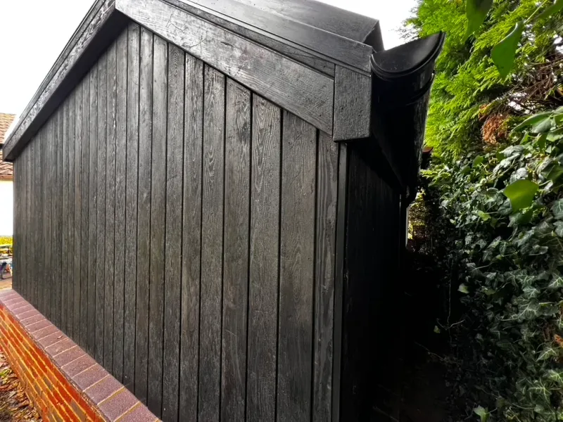 A garden building clad in charred black timber boards over a brick plinth, framed by greenery on a wet day.