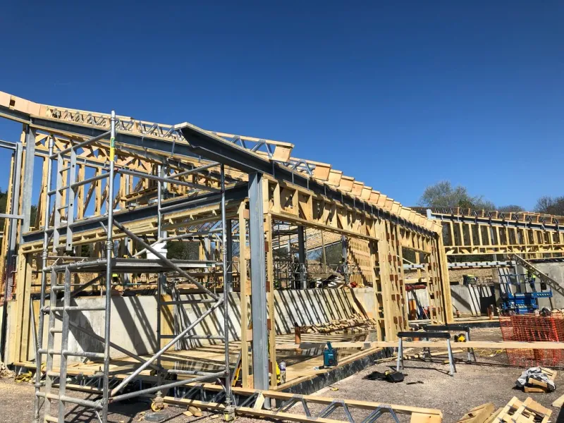 A timber frame with steel beams going up on site behind scaffolding, under a clear blue sky.