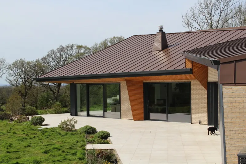 A completed contemporary new build with a brown standing-seam zinc roof, timber and brick elevations and full-height glazing onto a paved terrace.