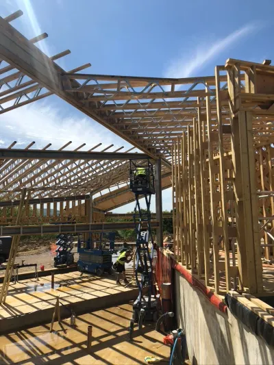 A timber-frame new build under construction, engineered roof trusses and stud walls rising over a concrete slab under a clear sky.