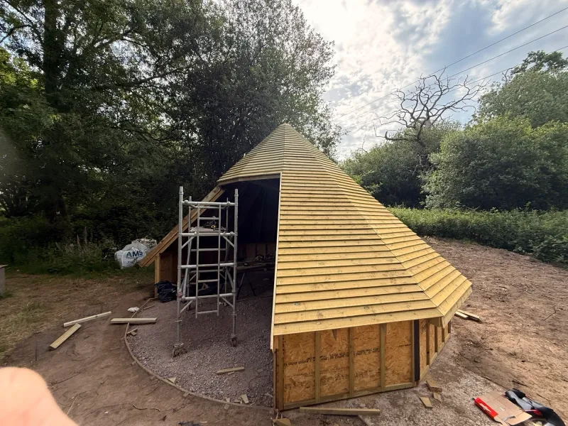 The ten-sided fire-pit hut during the build, its pointed roof boarded in timber, with a scaffold tower beside it in the woodland.